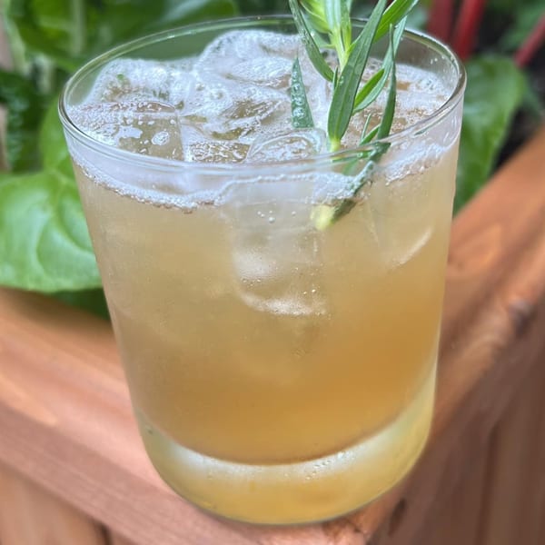 A refreshing golden drink with ice and a green herb garnish, set against a leafy green background.