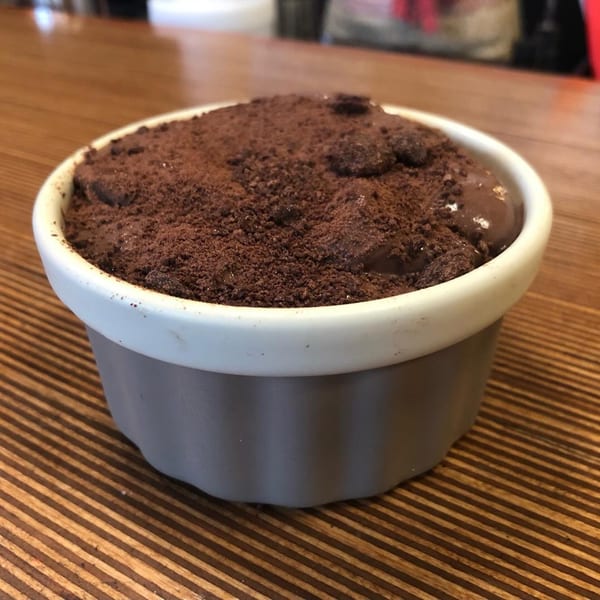 A chocolate dessert in a white and gray ramekin, topped with chocolate crumbles on a wooden table.