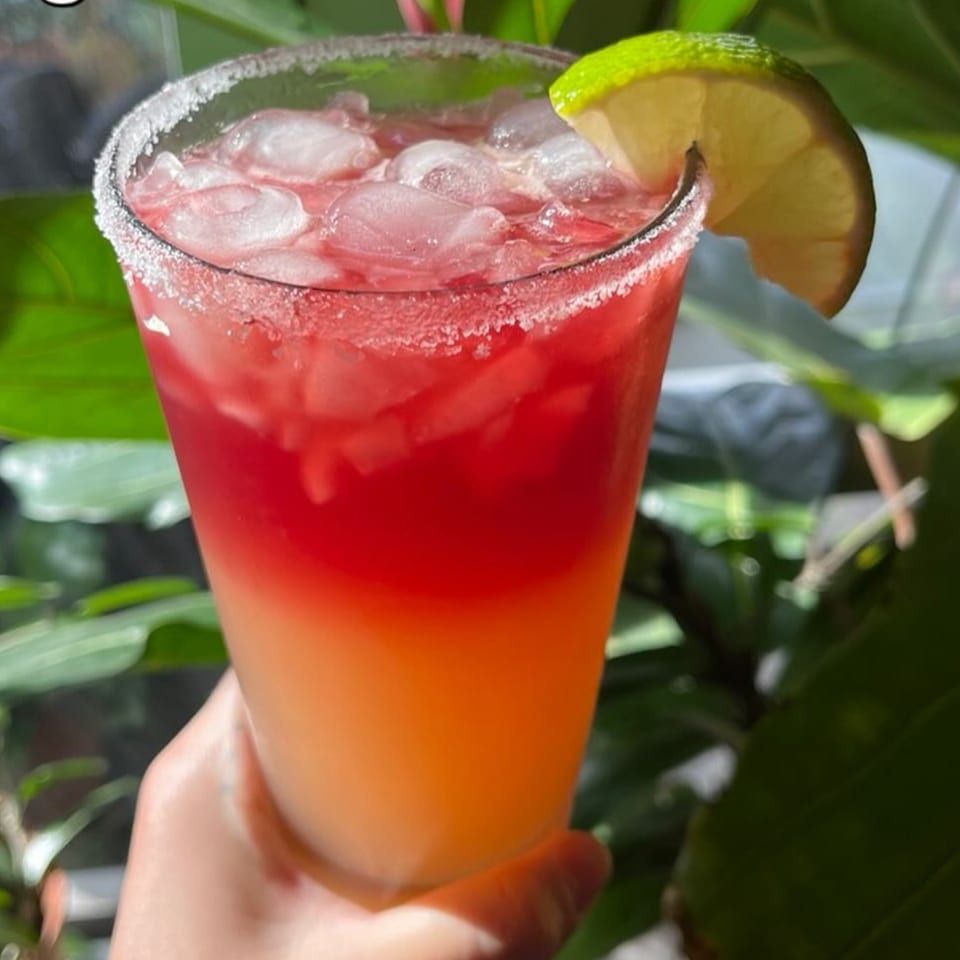 Hand holding a vibrant red and orange layered drink with ice, a salted rim, and a lime wedge.