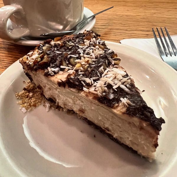 A slice of vegan cheesecake with chocolate, coconut, and nuts on a white plate with a fork.